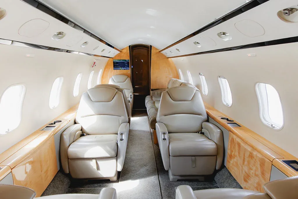 The interior cabin of a private jet with light beige leather seats and light wood paneling.