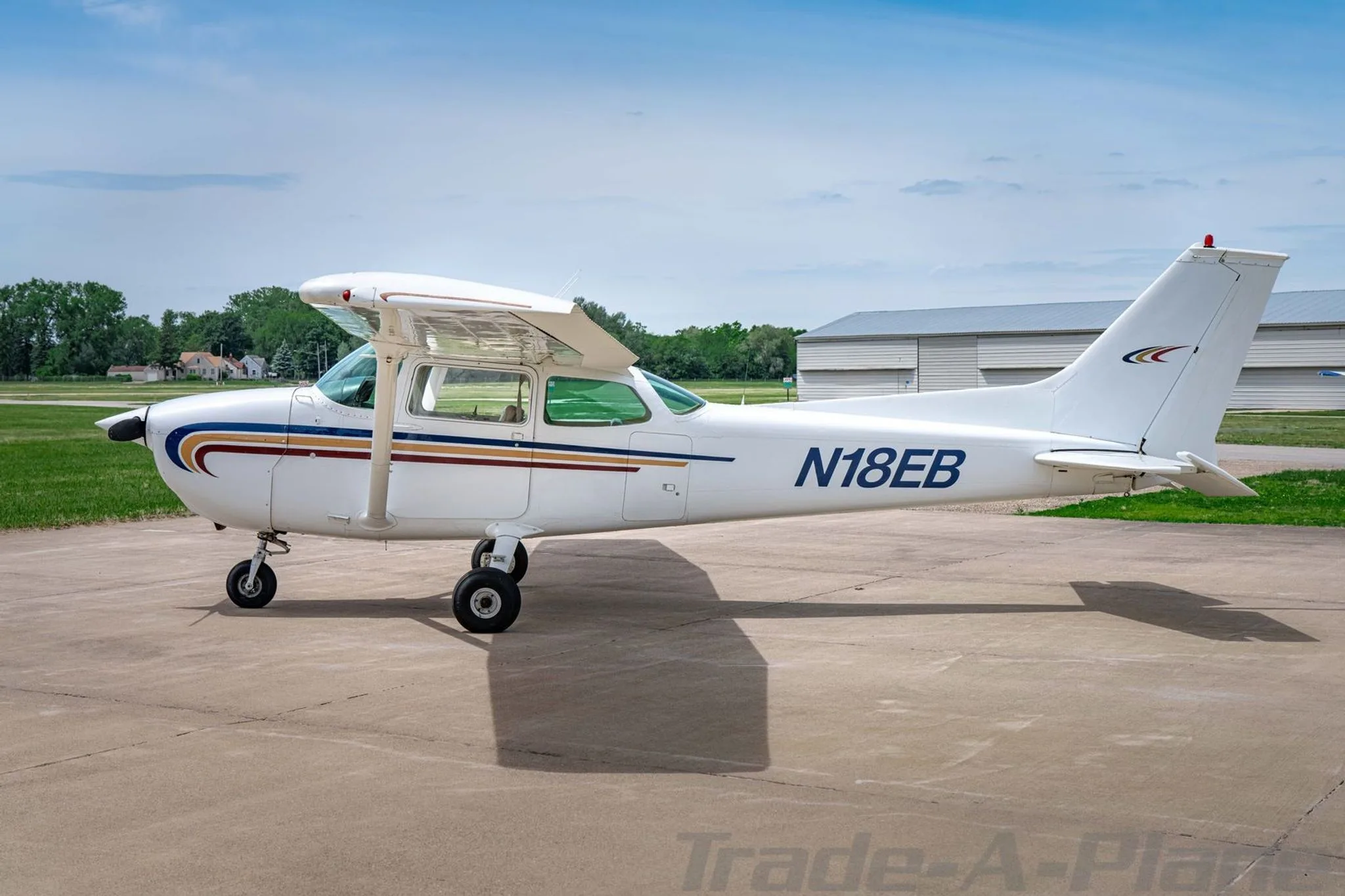 A white Cessna 172 with blue, gold, and red stripes on the fuselage, parked on an asphalt ramp, viewed from the left side.