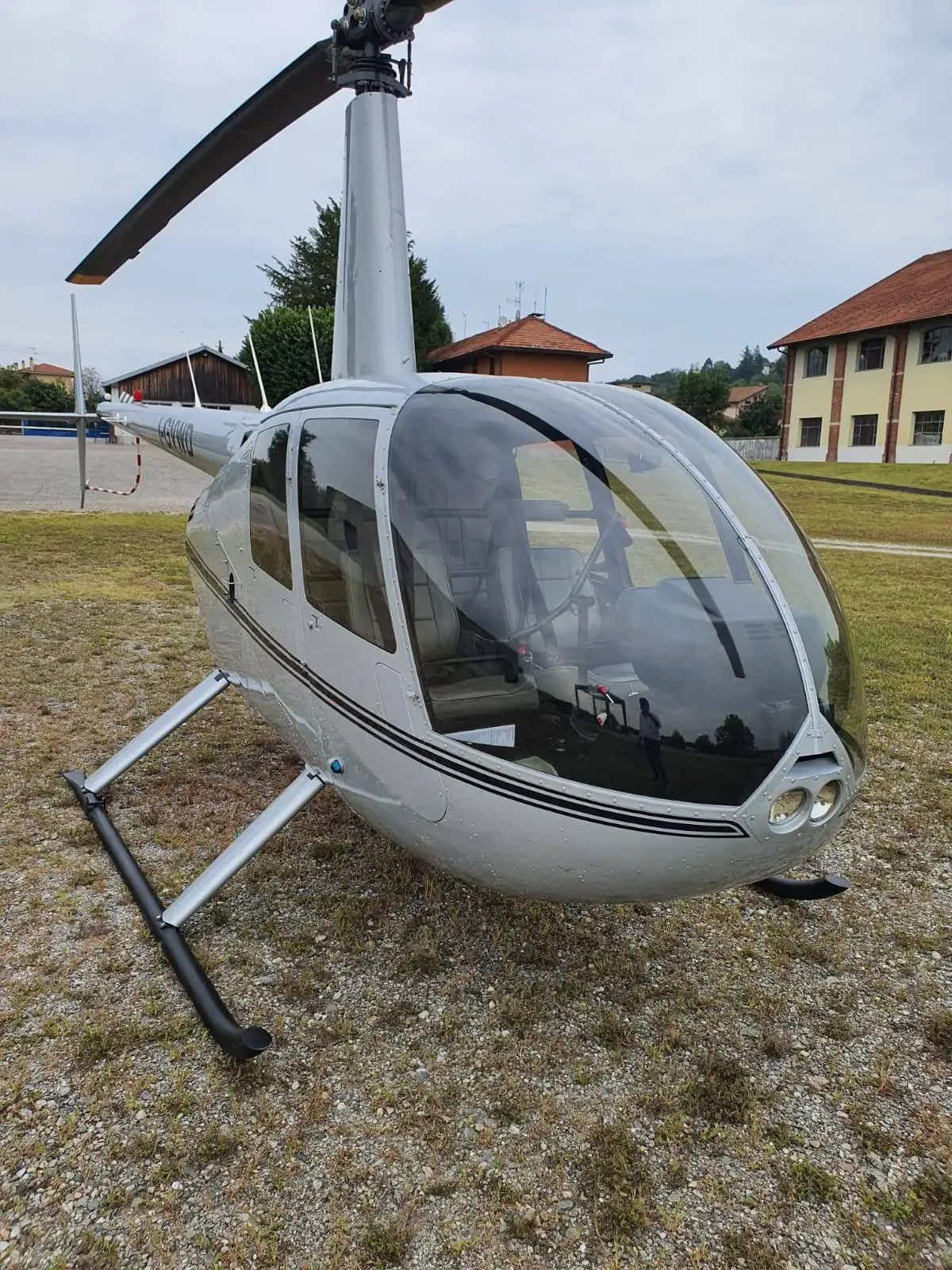A silver Robinson R44 helicopter parked on a grassy field with buildings in the background.