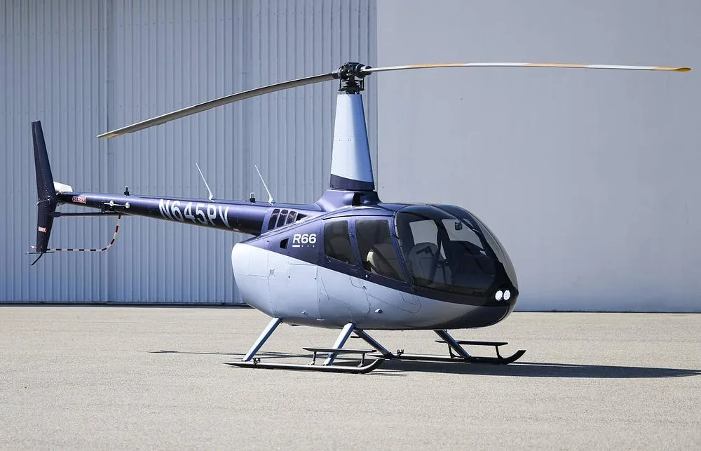 A blue and silver Robinson R66 helicopter (N645PV) is parked on an asphalt surface next to a white hangar.