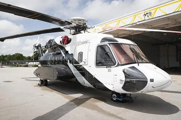 A white and black helicopter parked on an asphalt surface, viewed from the front-right, with a hangar in the background.