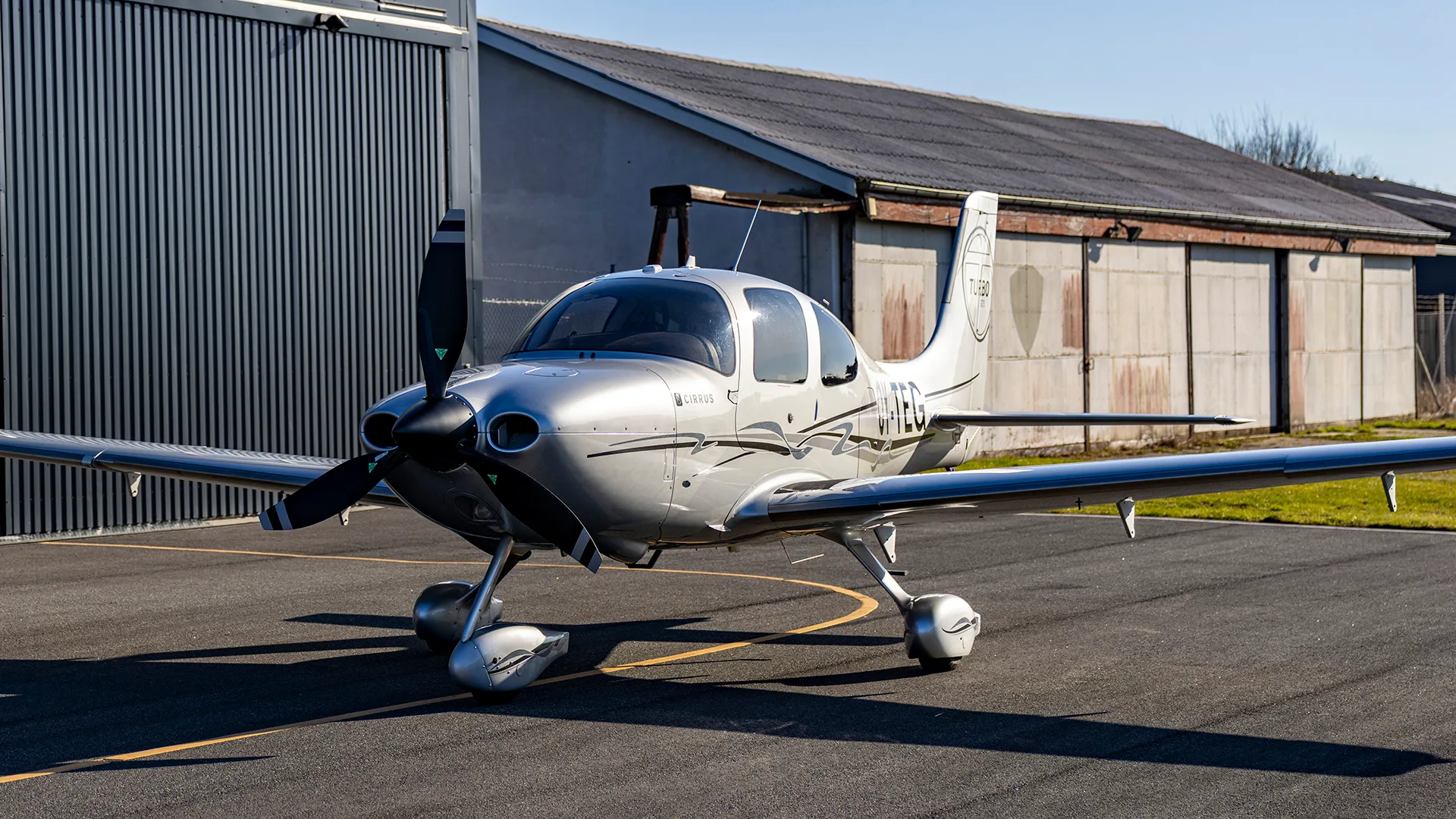 A silver Cirrus SR22T aircraft with registration T-GSO parked on an asphalt apron in front of a hangar.