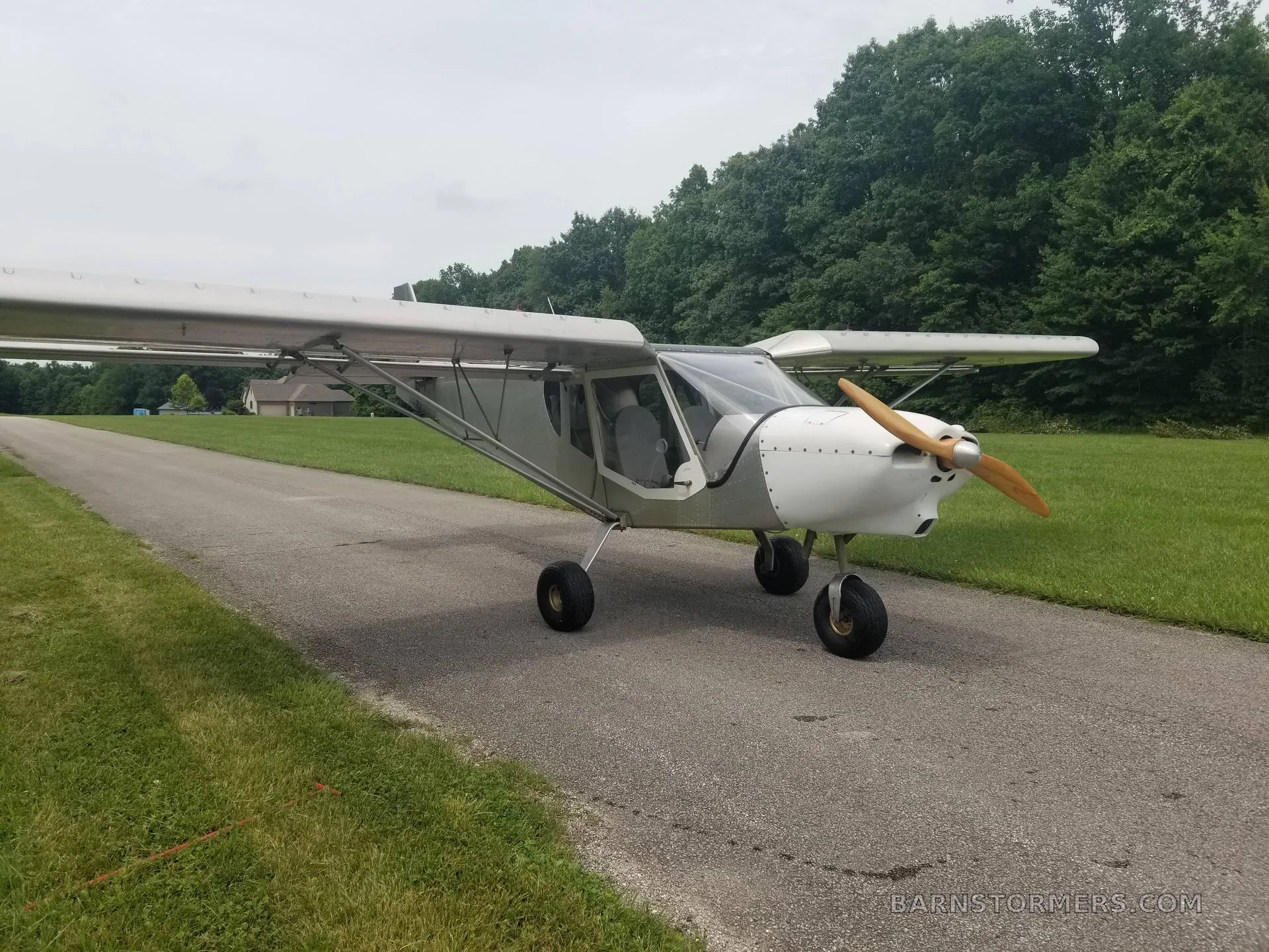 A light gray ultralight aircraft with a wooden propeller is parked on a paved path, with green grass and trees in the background.