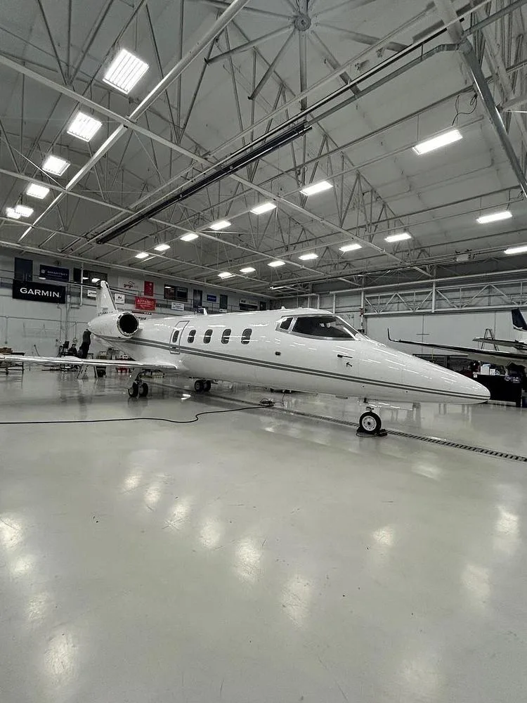 A white Learjet business jet with a grey stripe is parked in a brightly lit hangar.