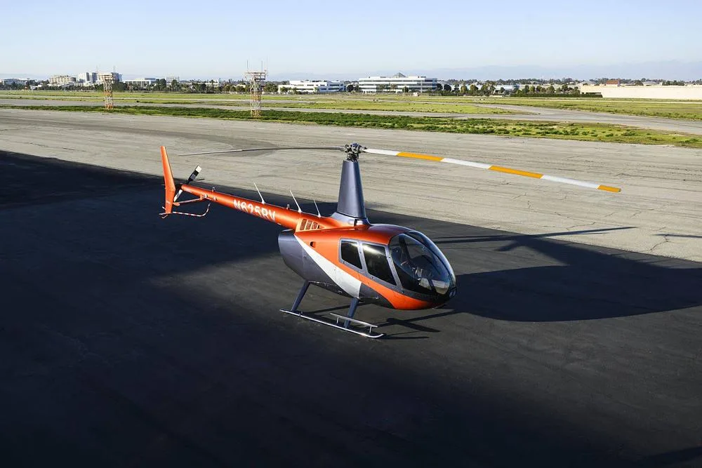 A two-tone orange and grey helicopter (N805RV) with white and orange rotor blades sits on an airfield under a clear sky.