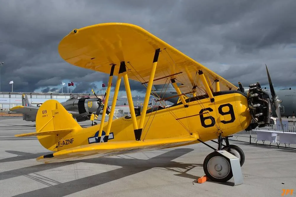 A bright yellow biplane with black '69' markings is parked on a tarmac under a cloudy sky, with other aircraft in the background.