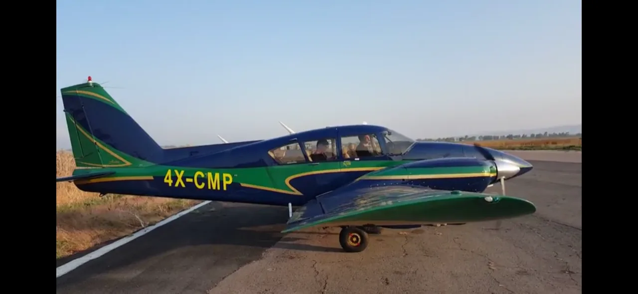 A dark blue and green single-engine propeller aircraft with registration 4X-CMP is parked on an asphalt surface with fields in the background.