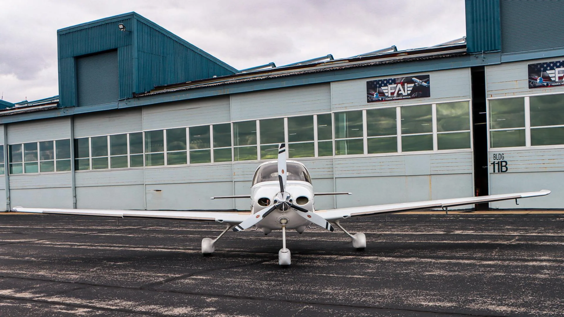 A white Cirrus aircraft with a three-bladed propeller is parked on a tarmac in front of a hangar.