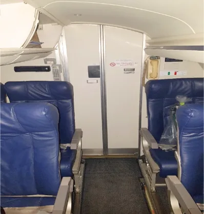 The aircraft interior shows blue passenger seats on either side of an aisle leading to a forward bulkhead.