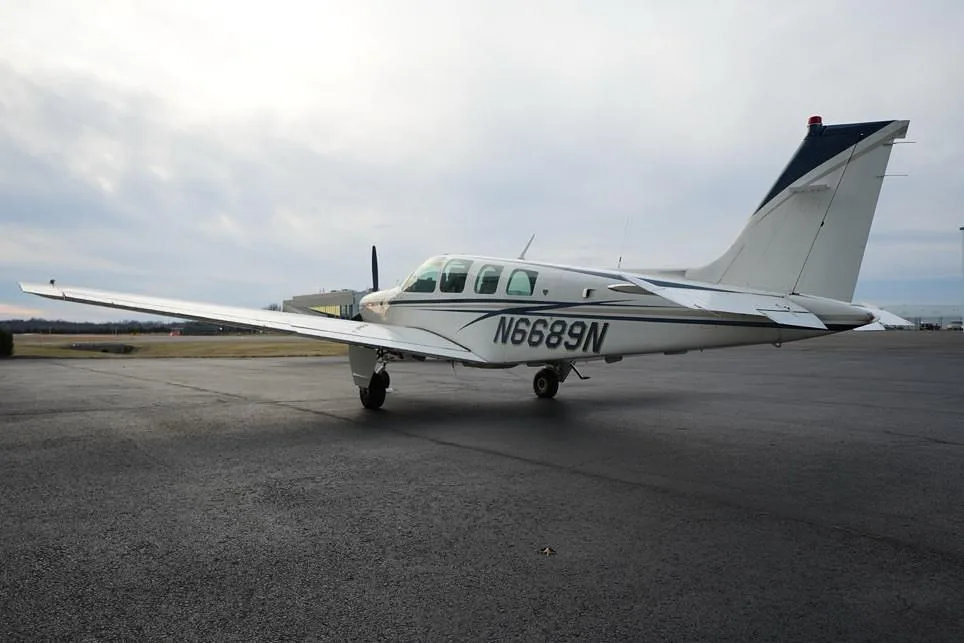A rear-side view of the white Beechcraft Bonanza with blue and gray stripes, registration N6689N, parked on an airport ramp.