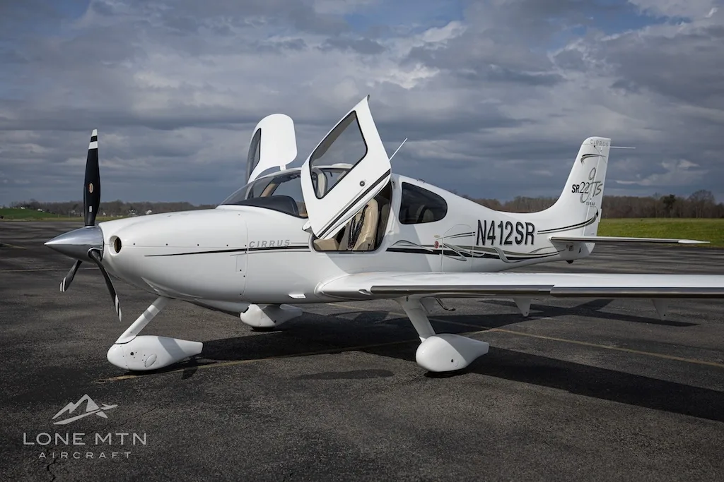 A white Cirrus SR22T aircraft with open doors is parked on an asphalt runway under a cloudy sky.