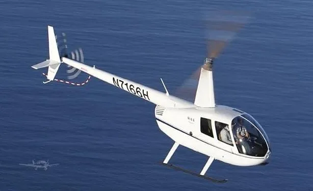 A white Robinson R44 helicopter with tail number N7166H flies over blue water.