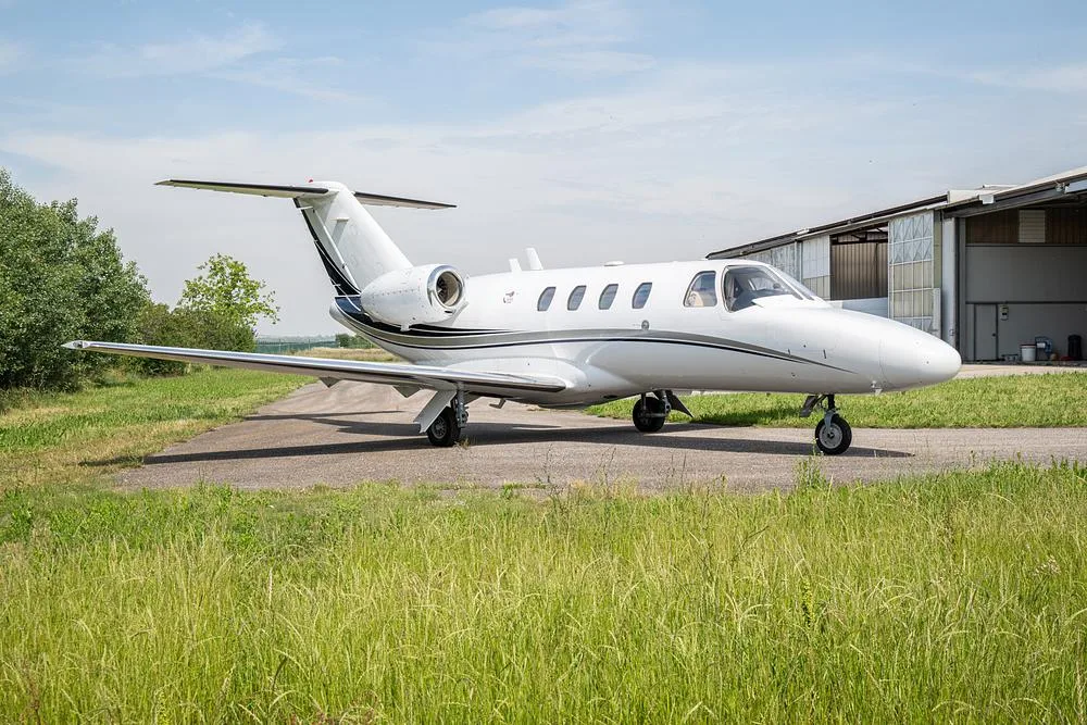 A white business jet with black and blue stripes parked on a tarmac next to a hangar on a sunny day.