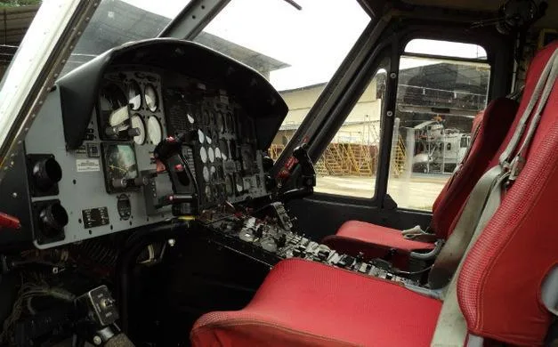 A view from inside a helicopter cockpit showing the instrument panel, controls, and two red pilot seats.