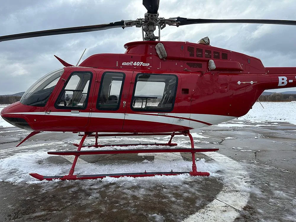A close-up side view of a red Bell 407GXP helicopter with white stripes, parked on a partially snowy ground.