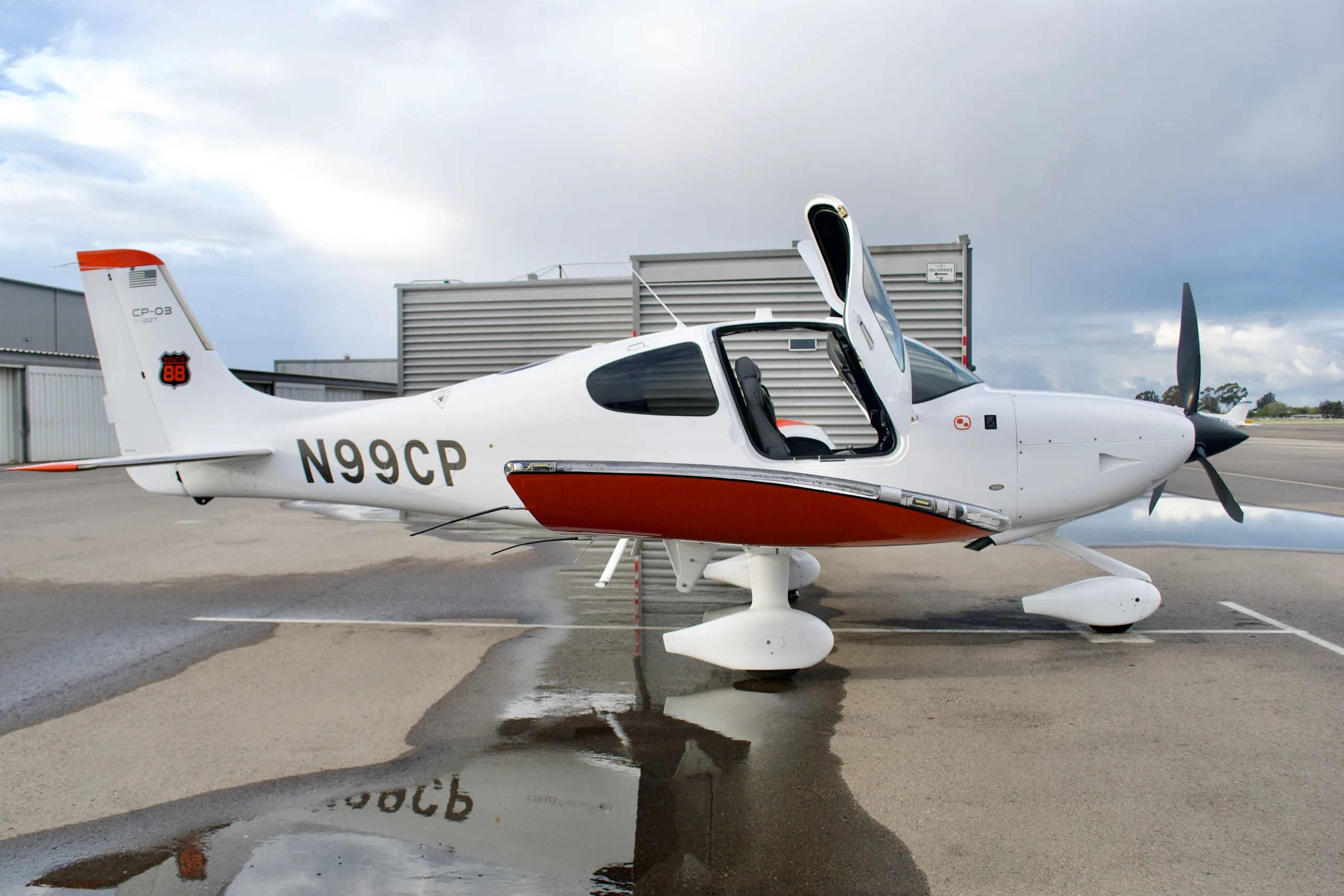 A white Cirrus SR22T aircraft with orange accents and registration N99CP, with its door open, parked on a wet tarmac.
