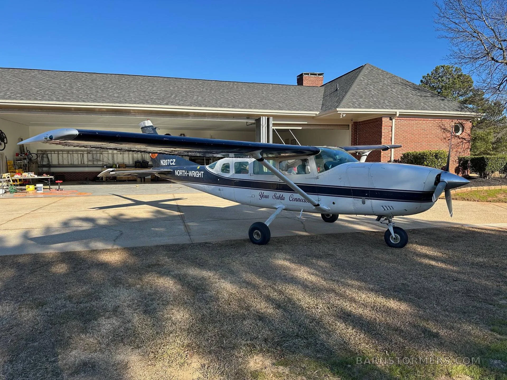A white and dark blue Cessna 207 aircraft, N207CZ, with "NORTH-WRIGHT" text, parked on a concrete driveway in front of a garage.