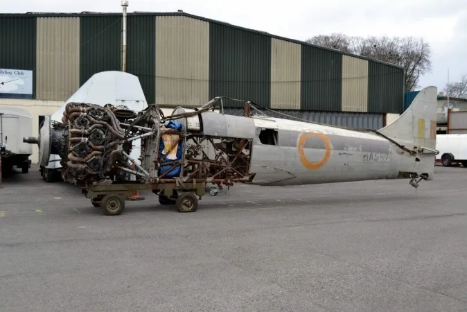 A disassembled aircraft fuselage and radial engine on a cart, parked outdoors next to a hangar.