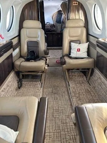 The interior cabin of a private aircraft features tan leather seats and patterned carpet, looking towards the rear.