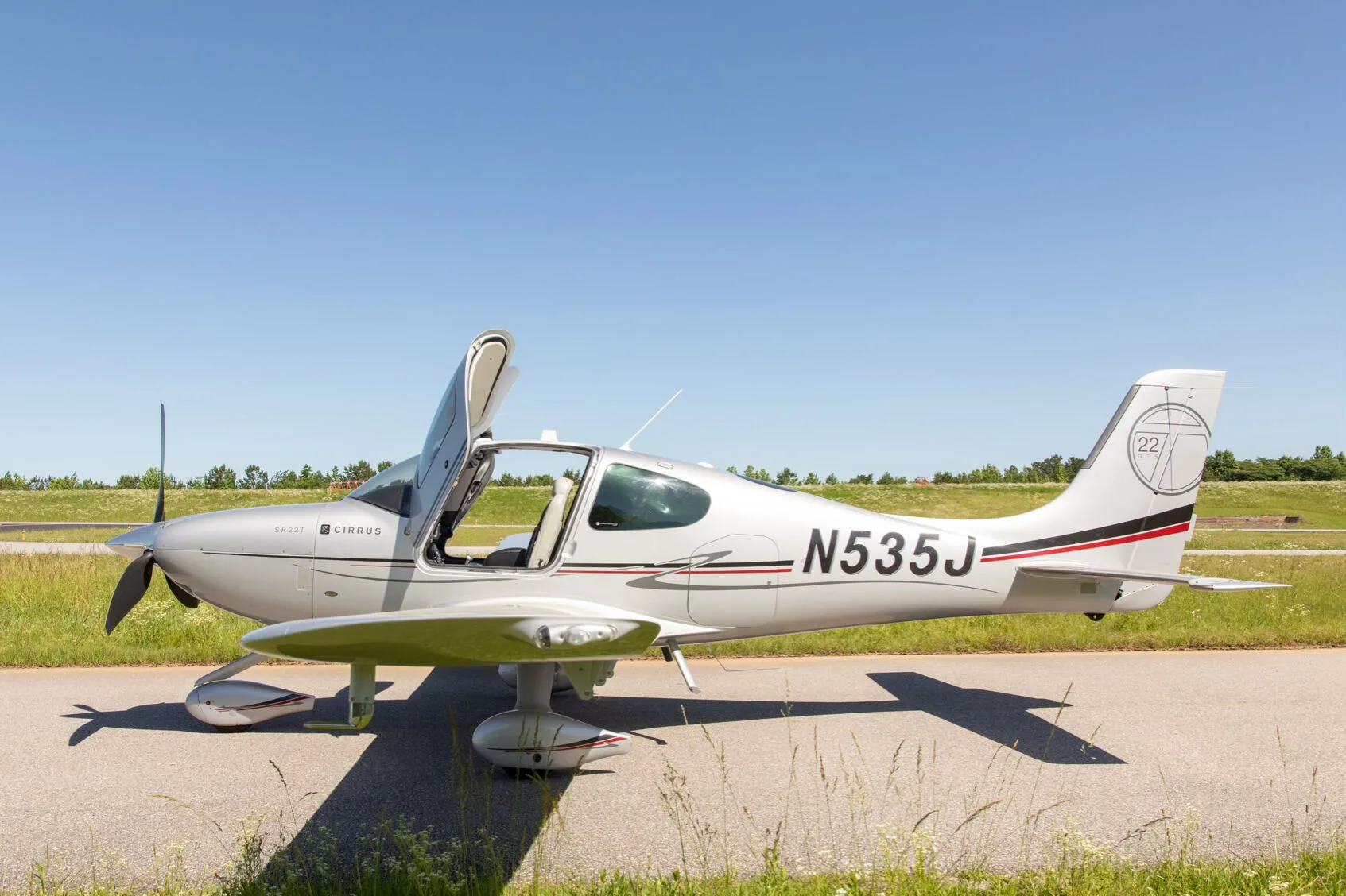 A silver Cirrus SR22T aircraft with registration N535J is parked with its doors open on an asphalt strip next to green grass under a clear blue sky.