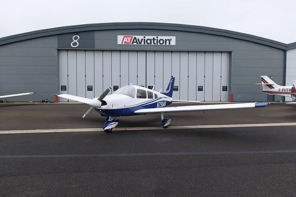 A white and blue Piper Cherokee aircraft, registration N75048, parked on an airfield in front of an AT Aviation hangar.