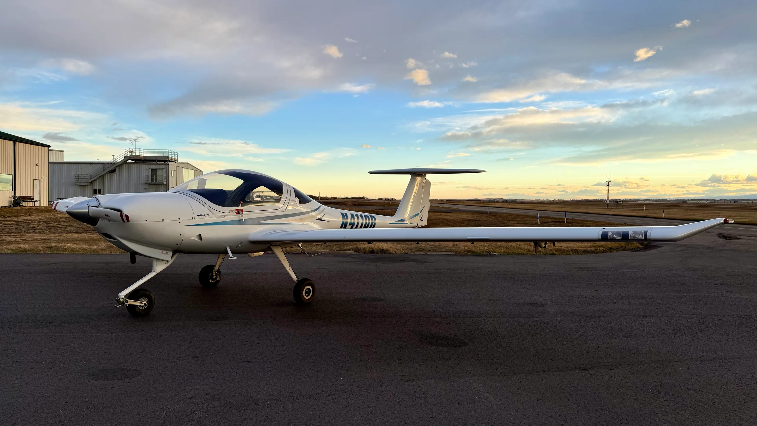 A white Diamond DA20-C1 Eclipse aircraft with blue stripes, registration N4110R, parked on an airfield with hangars in the background.