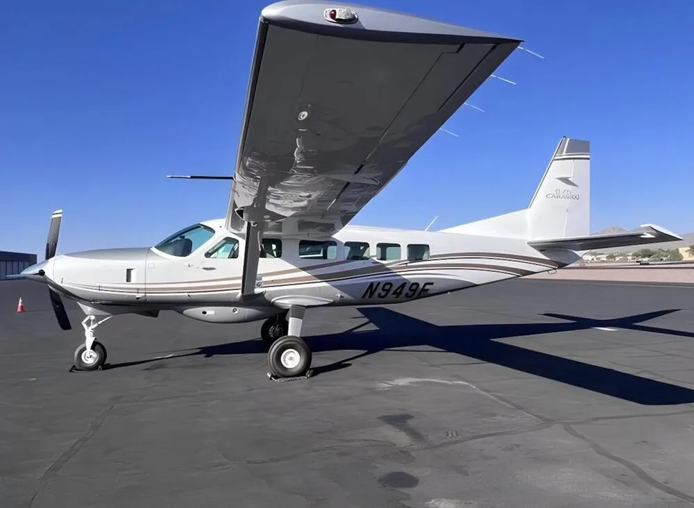 A silver Cessna Caravan N949F with brown stripes parked on an asphalt tarmac, viewed from the front-right.