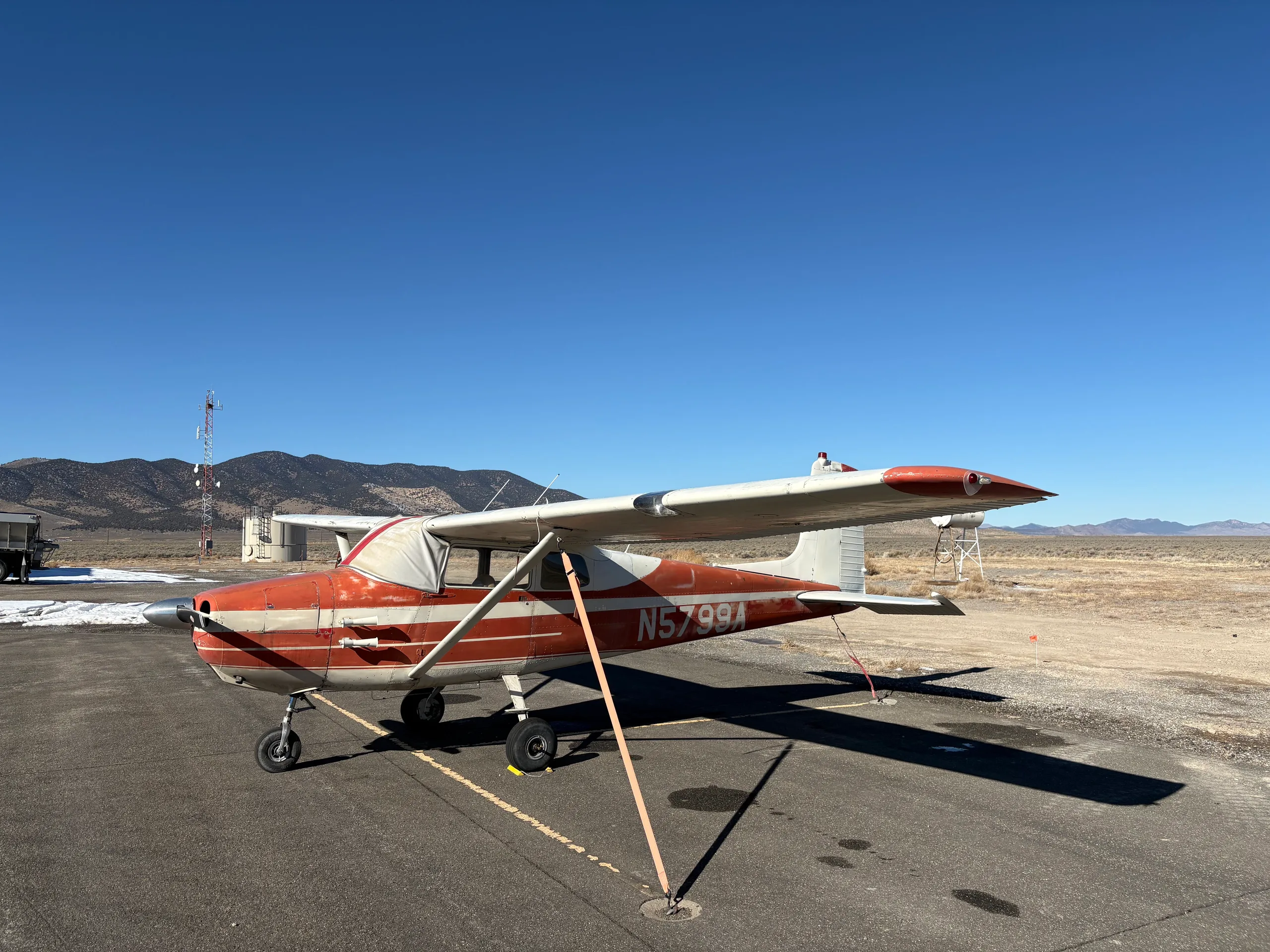 A red and white single-engine high-wing aircraft, N5799A, tied down on an asphalt ramp with mountains in the background.