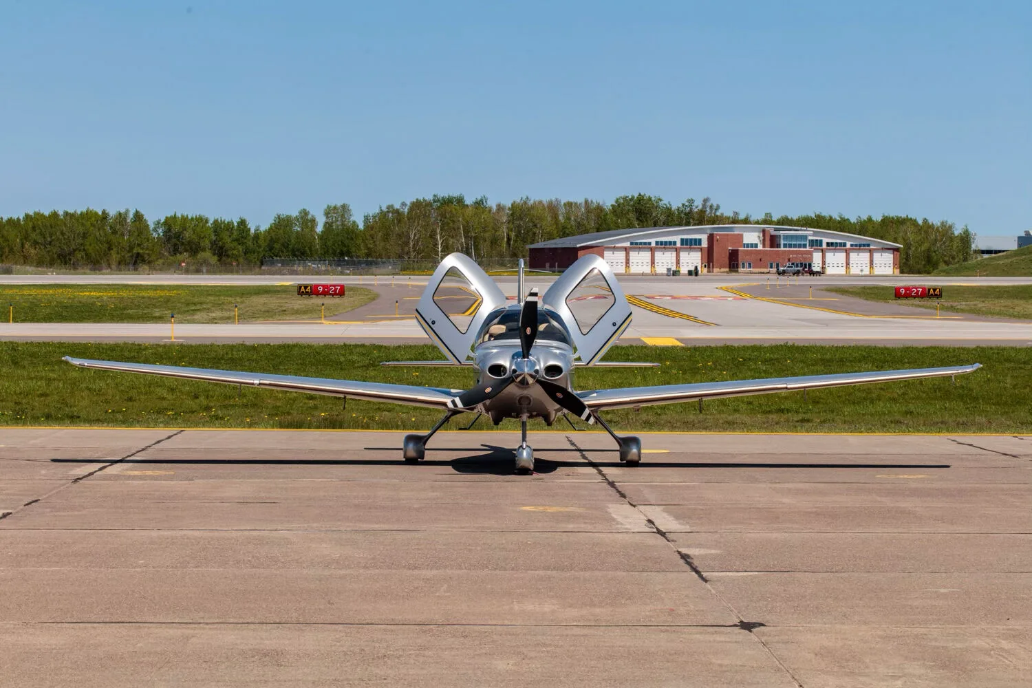 A silver Cirrus SR22T aircraft with its gull-wing doors open, parked on an airfield with hangars in the background.