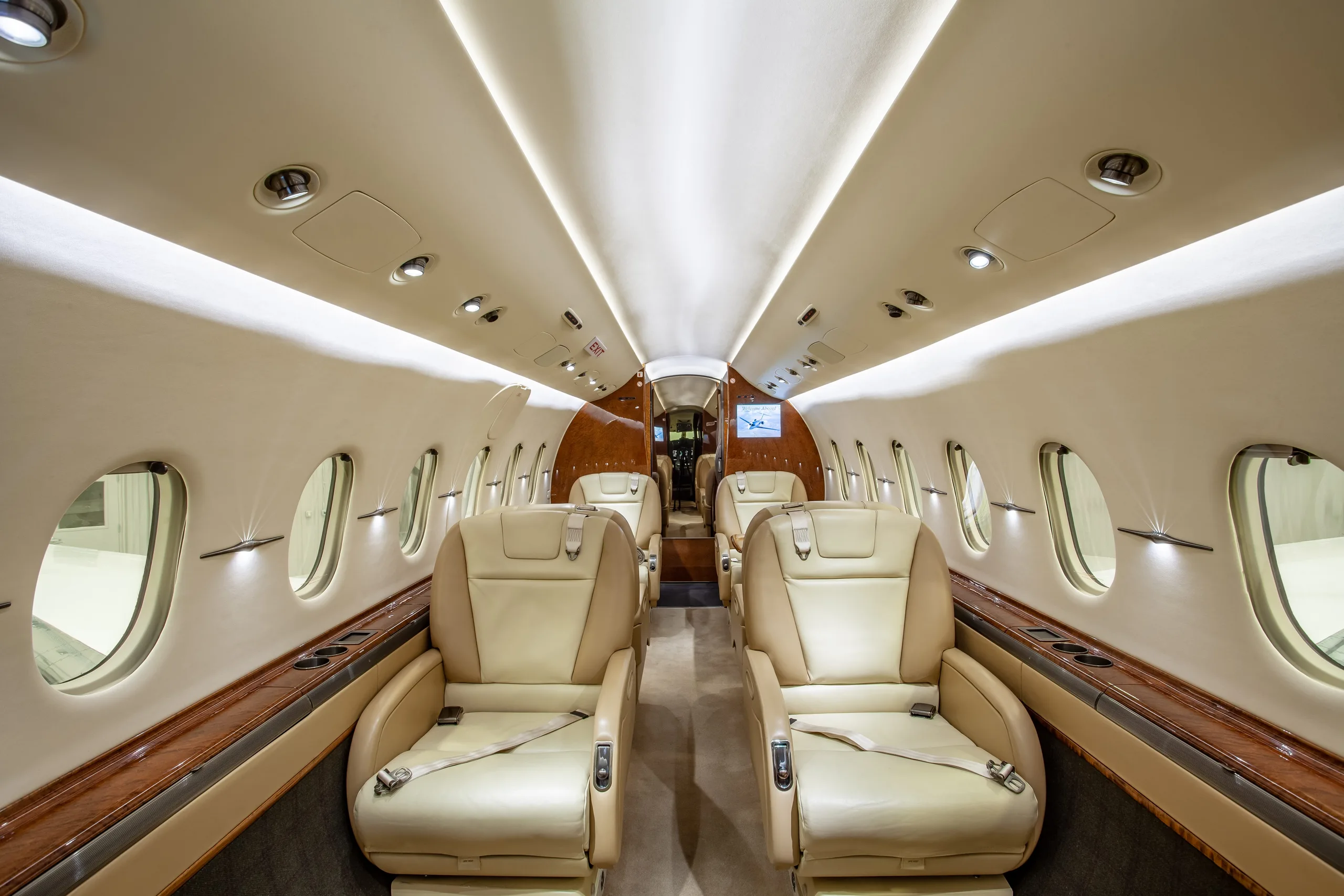 A wide view of a private jet's interior cabin featuring beige leather seats, large windows, and wood trim.
