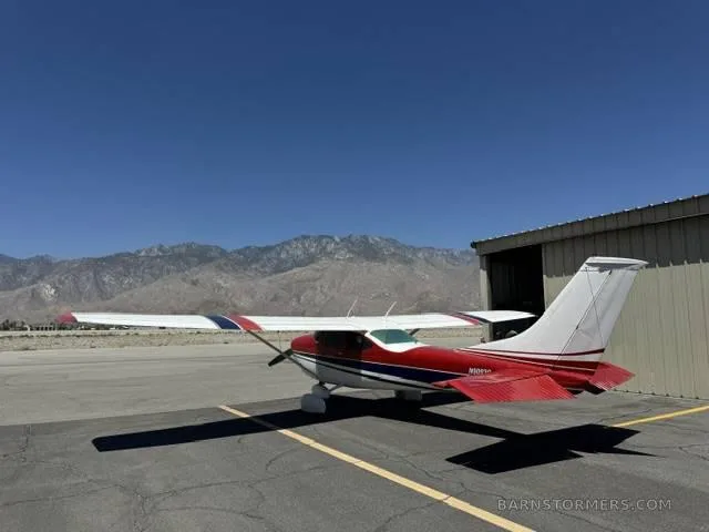 A red, white, and blue single-engine propeller aircraft parked on an asphalt ramp with mountains in the background.