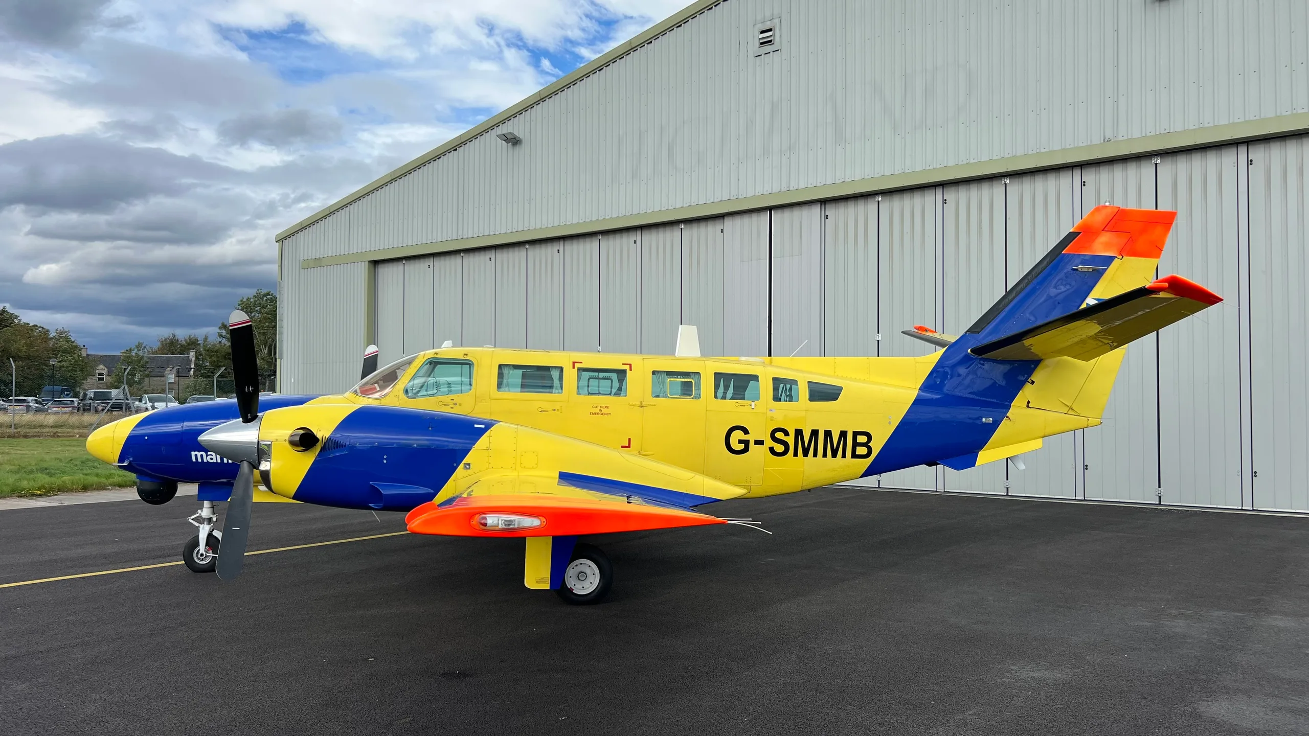 A yellow and blue twin-engine propeller aircraft, registration G-SMMB, is parked on a tarmac next to a large white hangar under a cloudy sky.