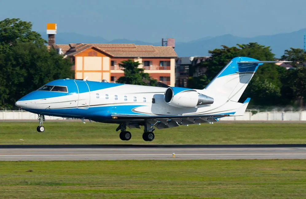 A blue and white Bombardier Challenger 604 private jet is captured mid-air during landing or takeoff.