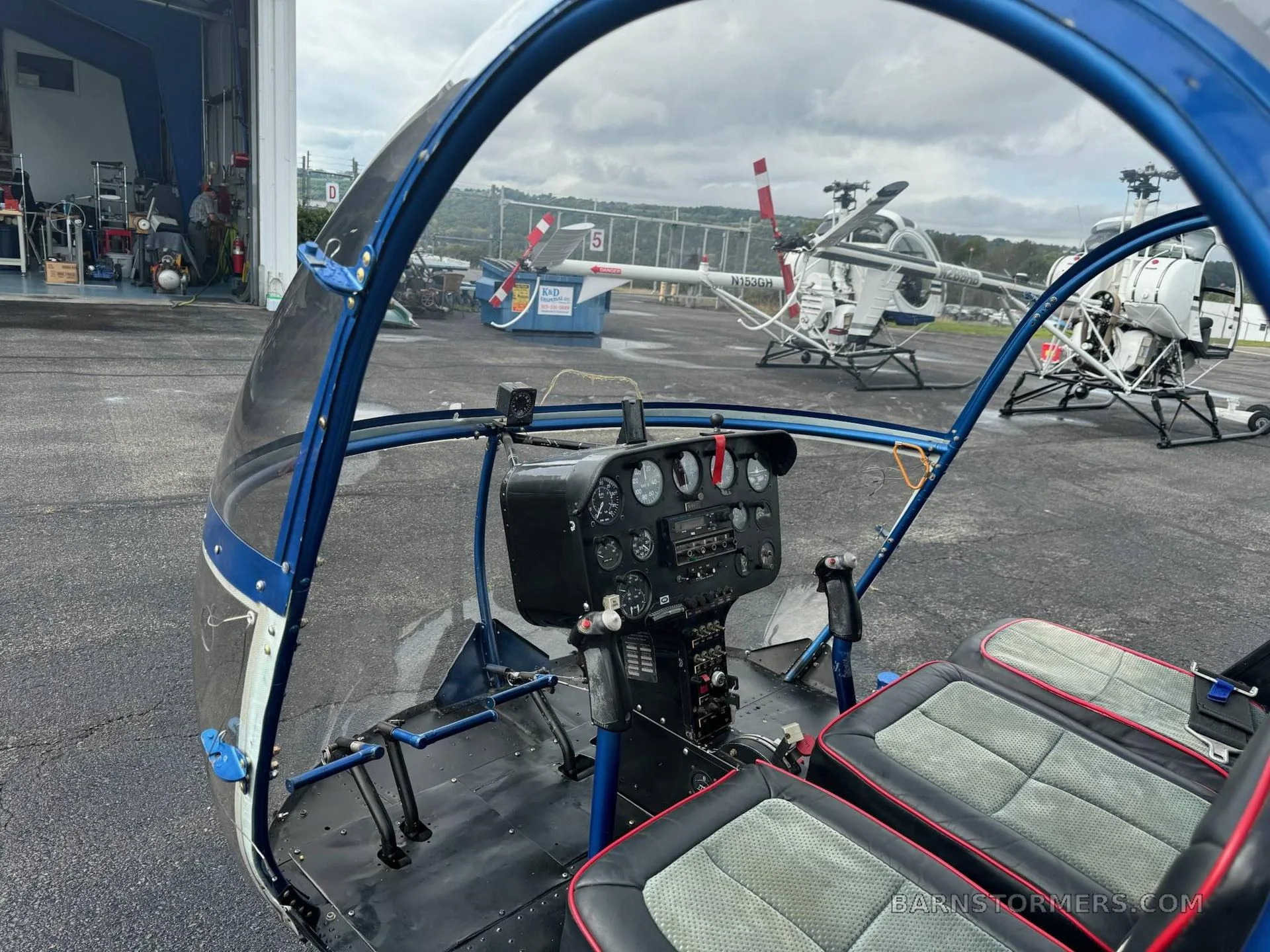 The cockpit of a blue and black helicopter, showing the instrument panel and seats, with other helicopters visible outside.
