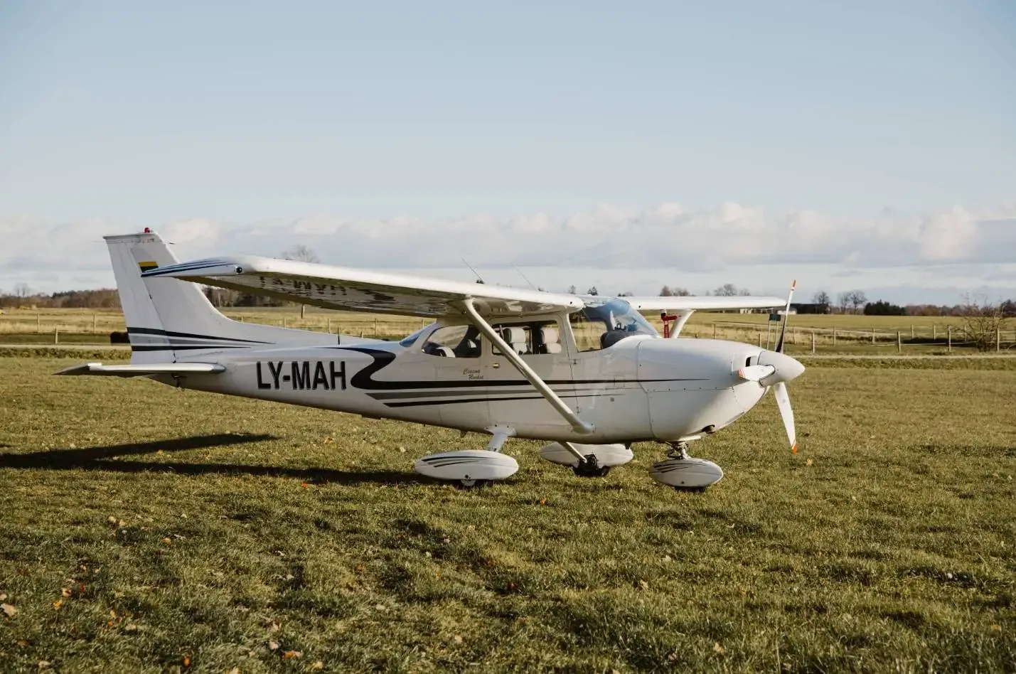 A white Cessna 172 with black and grey stripes, registration LY-MAH, parked on a grassy field under a clear sky.