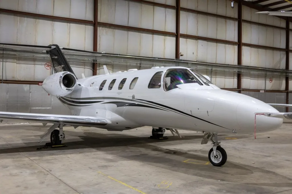 A white Cessna Citation M2 business jet with black and grey stripes is parked in a hangar, viewed from the front-right.