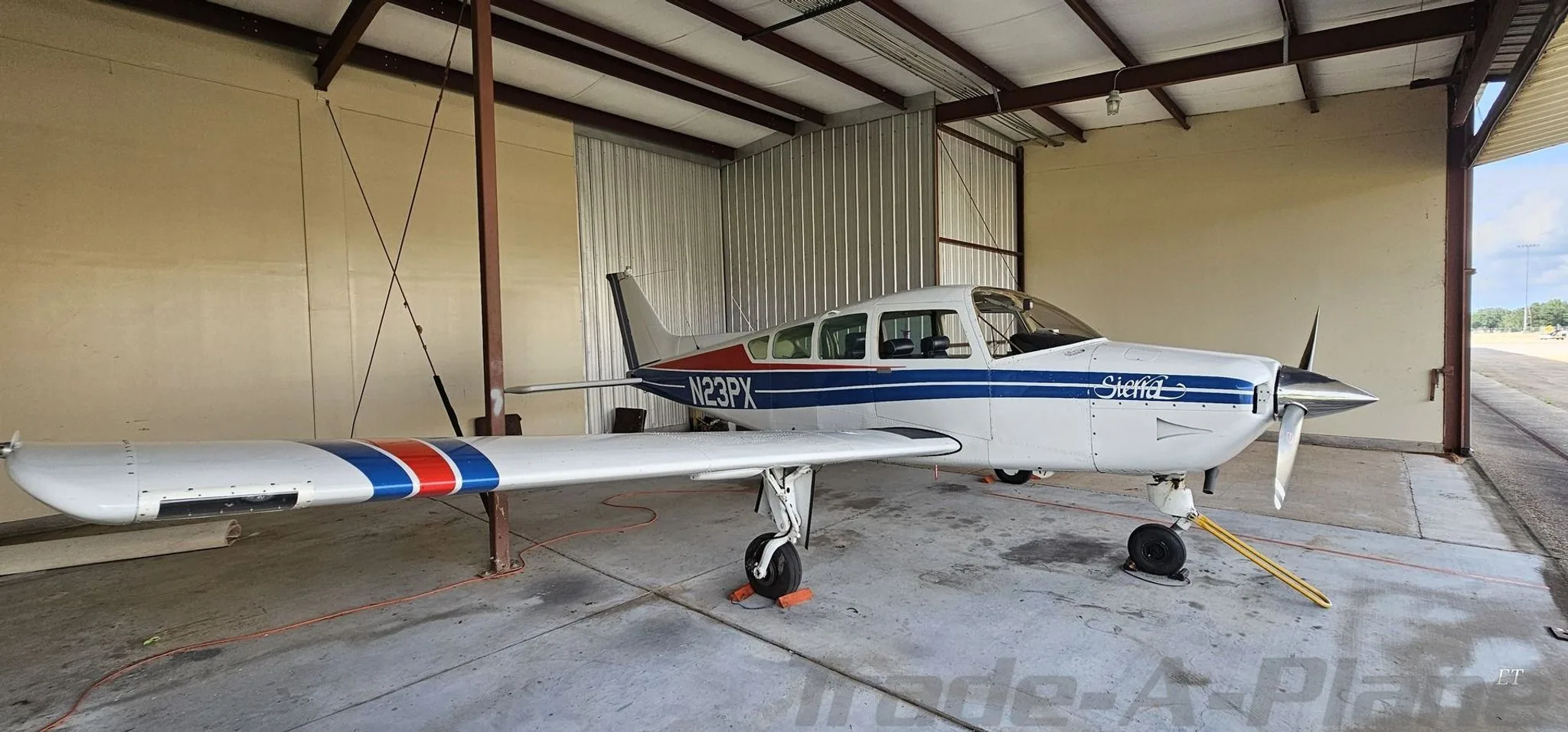A white Beechcraft Sierra N23PX with blue and red stripes parked in a hangar, viewed from the front right.