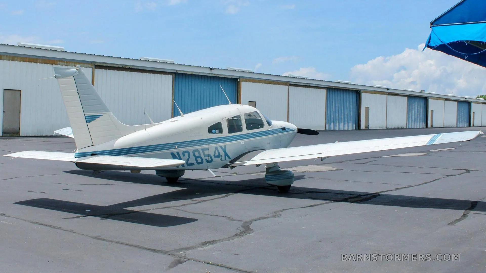 A white single-engine aircraft with blue stripes, registration N2854X, parked on an airfield with hangars in the background.