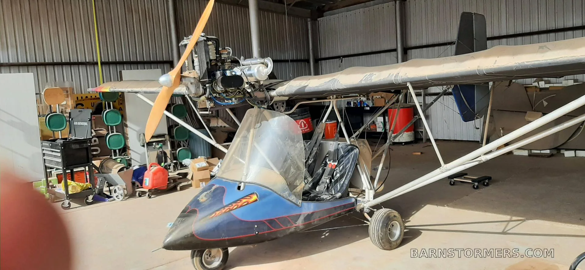 A blue and black ultralight aircraft with an exposed engine and propeller, parked inside a dusty hangar.