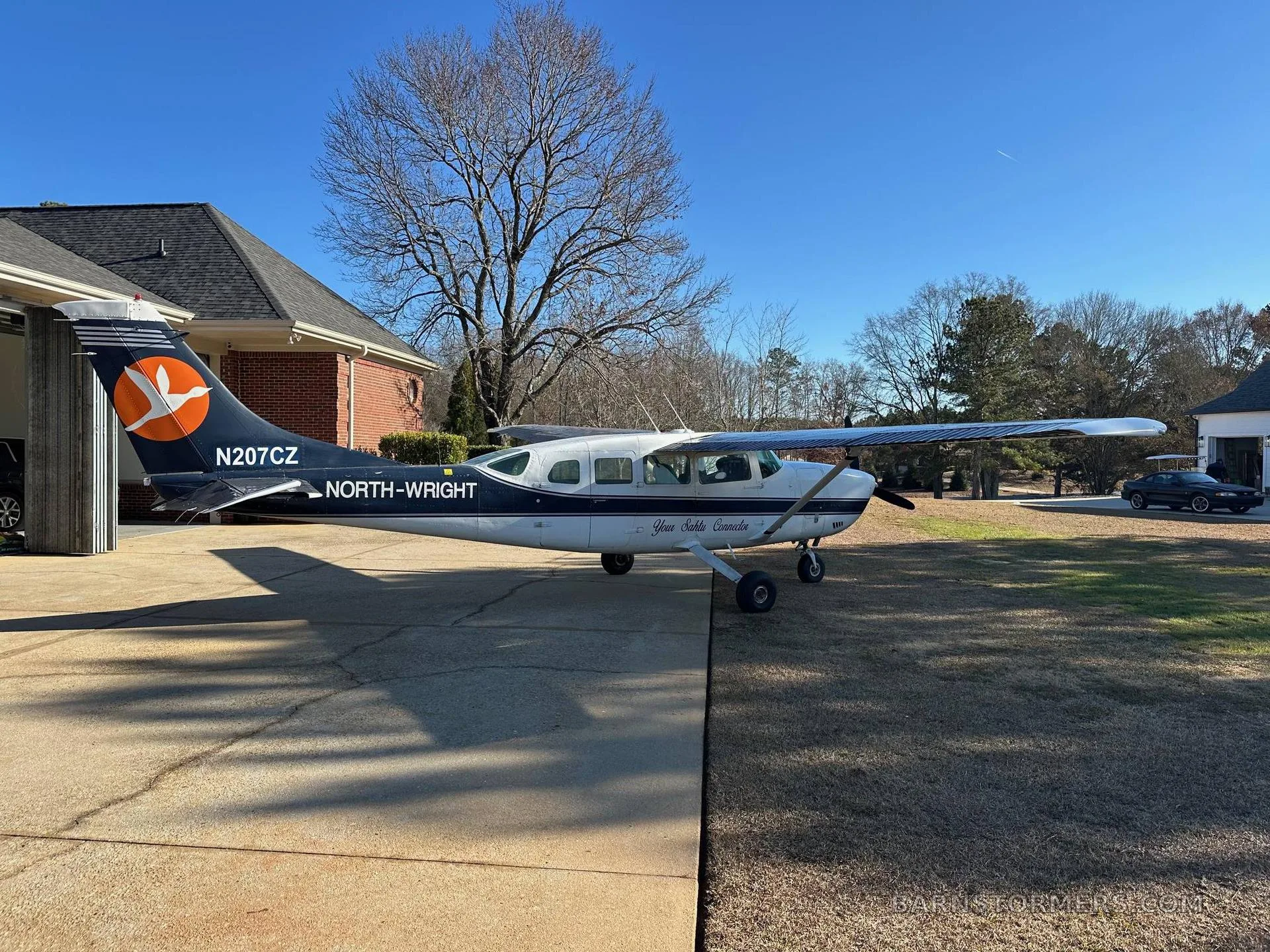 A white and dark blue Cessna 207 aircraft, N207CZ, with an orange bird logo on its tail, parked on a concrete driveway next to a house.