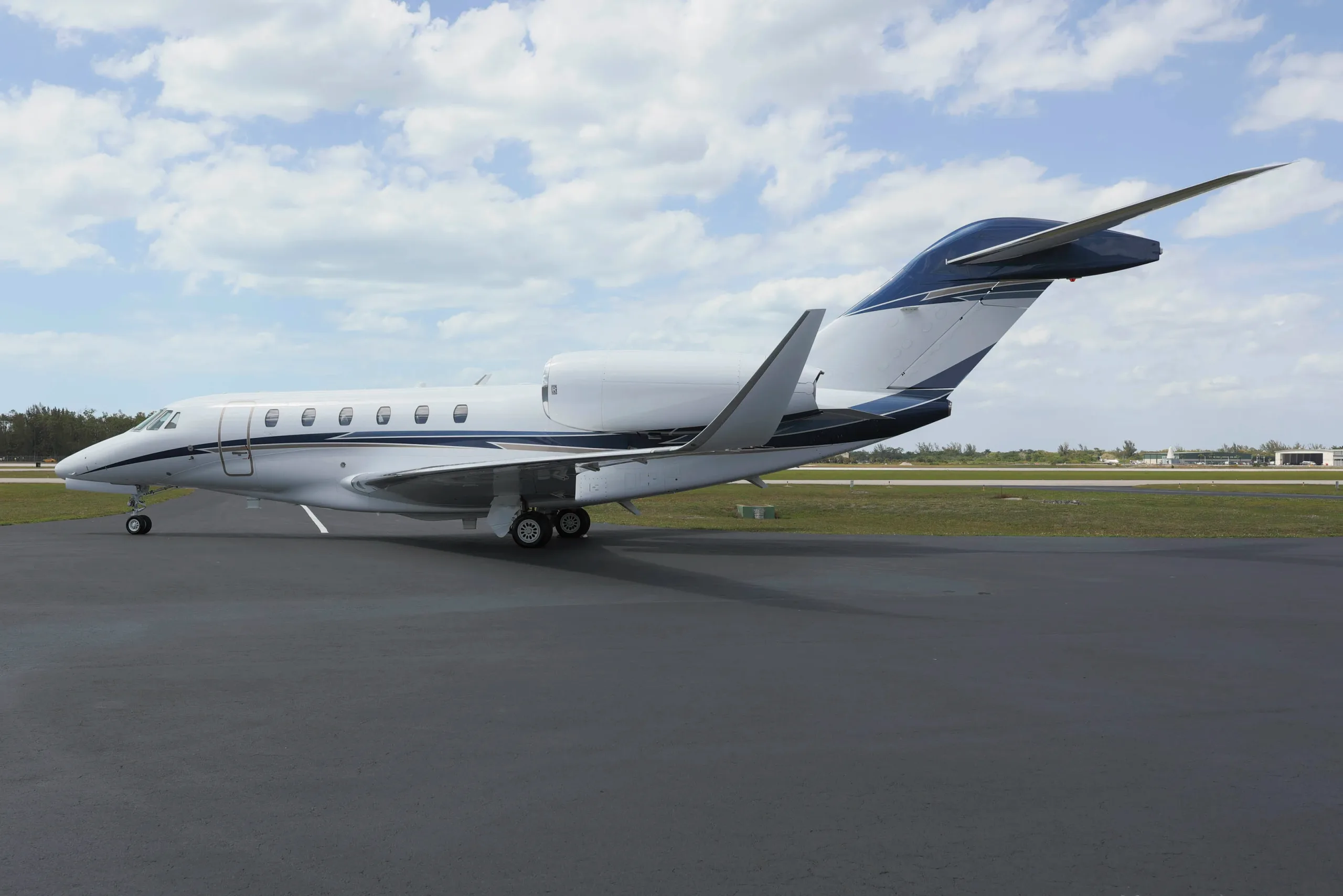 A white business jet with blue and grey stripes on the tail and fuselage is parked on an asphalt tarmac with a grassy field and distant buildings.