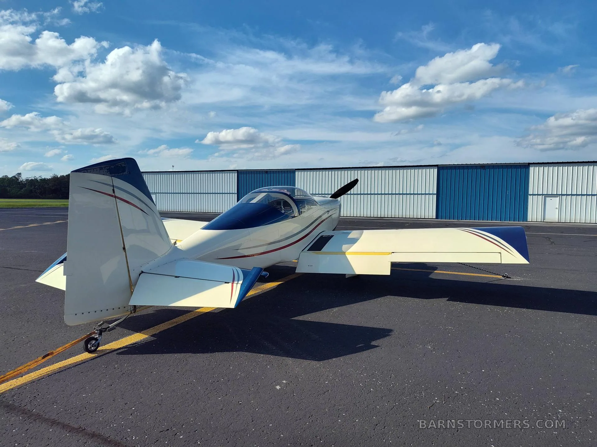 A cream and blue single-engine aircraft with red and grey stripes, viewed from the rear-left, parked on an asphalt tarmac.