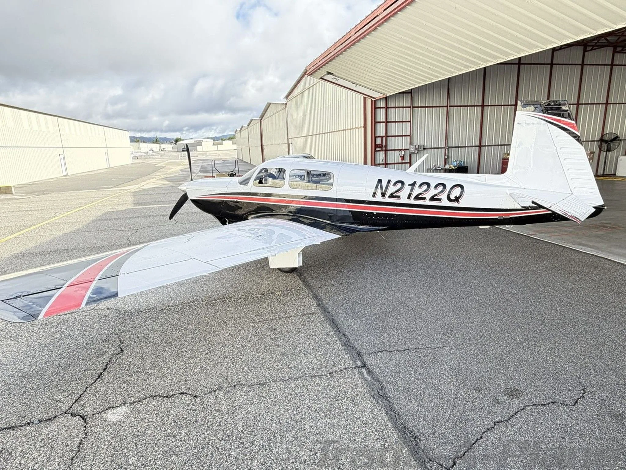 A white and black Mooney M20K aircraft with red stripes, registration N2122Q, parked on an tarmac next to a hangar.