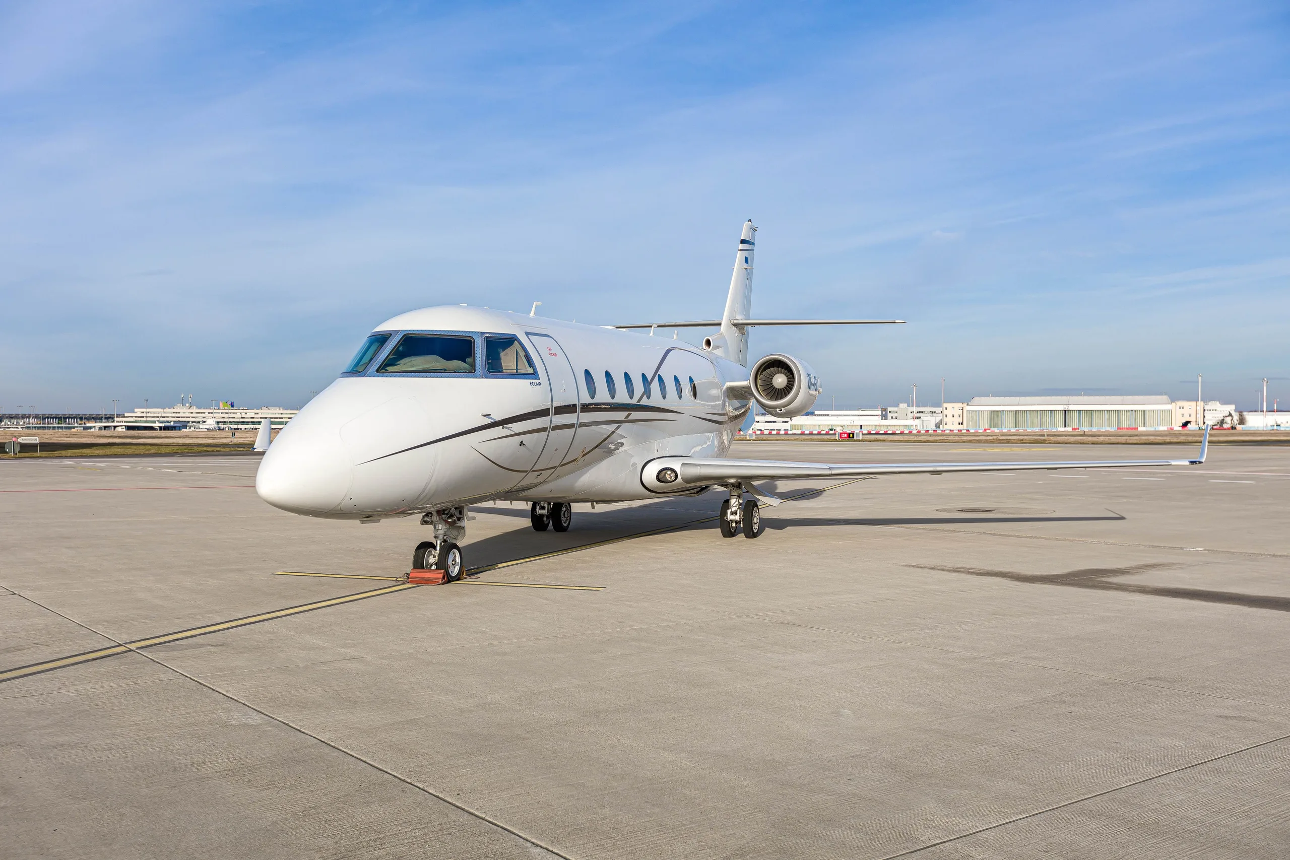 A white Gulfstream G200 business jet with brown accent stripes parked on an airport tarmac, viewed from the front-right.
