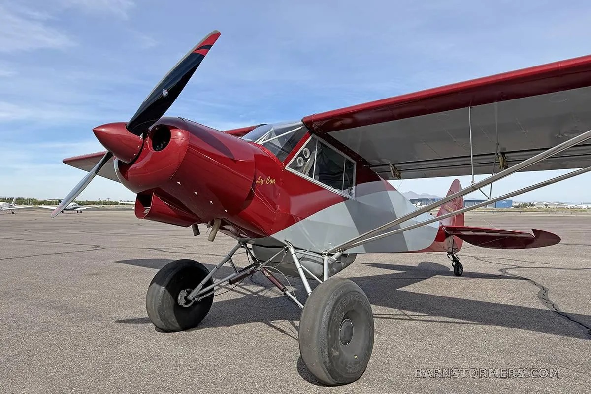 A red and grey high-wing aircraft with large tires, seen from a low front-right angle on an airfield.