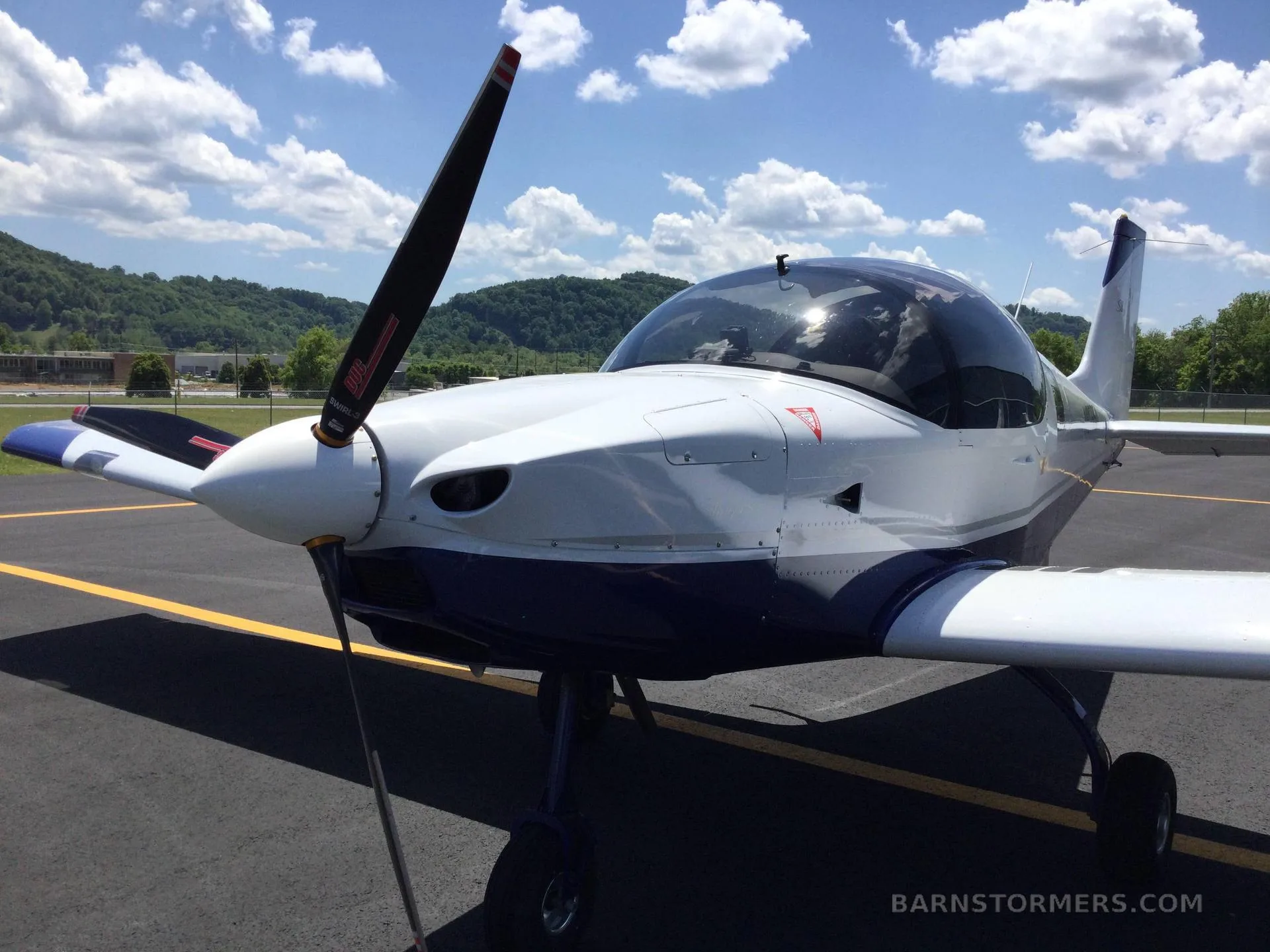 A white and blue Sling aircraft is parked on an asphalt runway under a partly cloudy sky, viewed from the front-right.