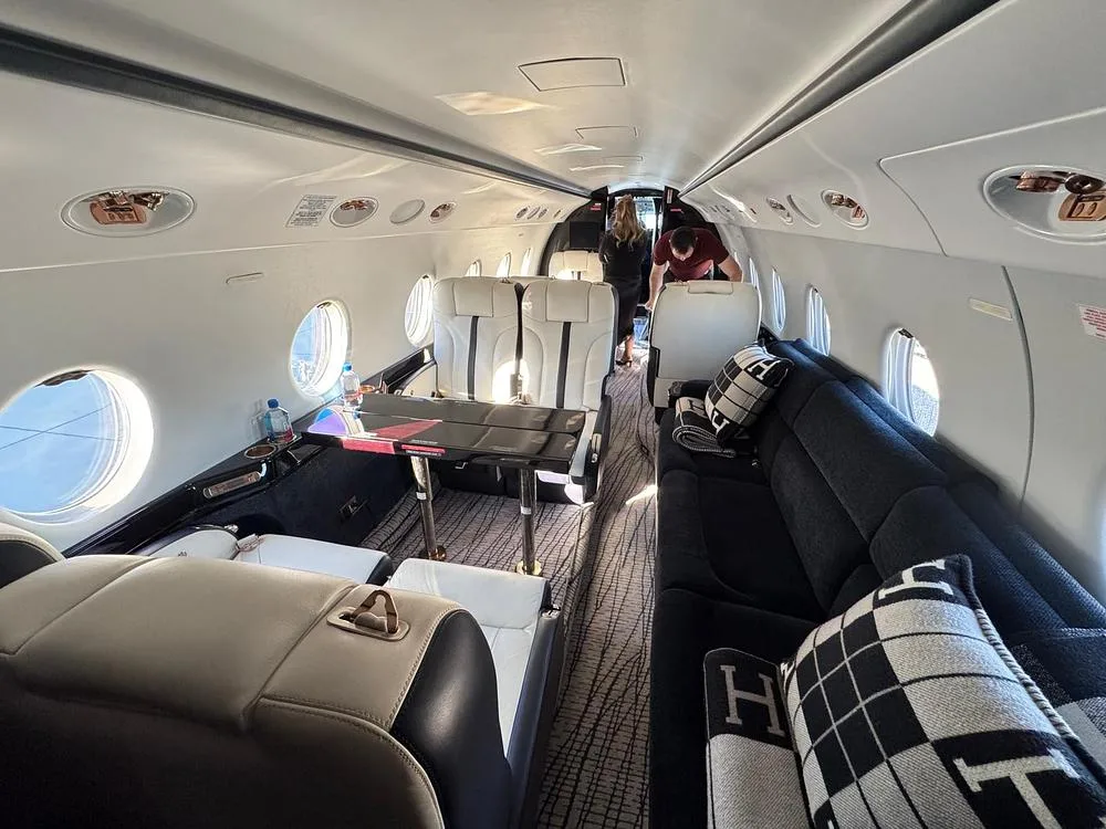The spacious interior cabin of a private jet with cream and black seating, a black couch with patterned pillows, and a table setup.