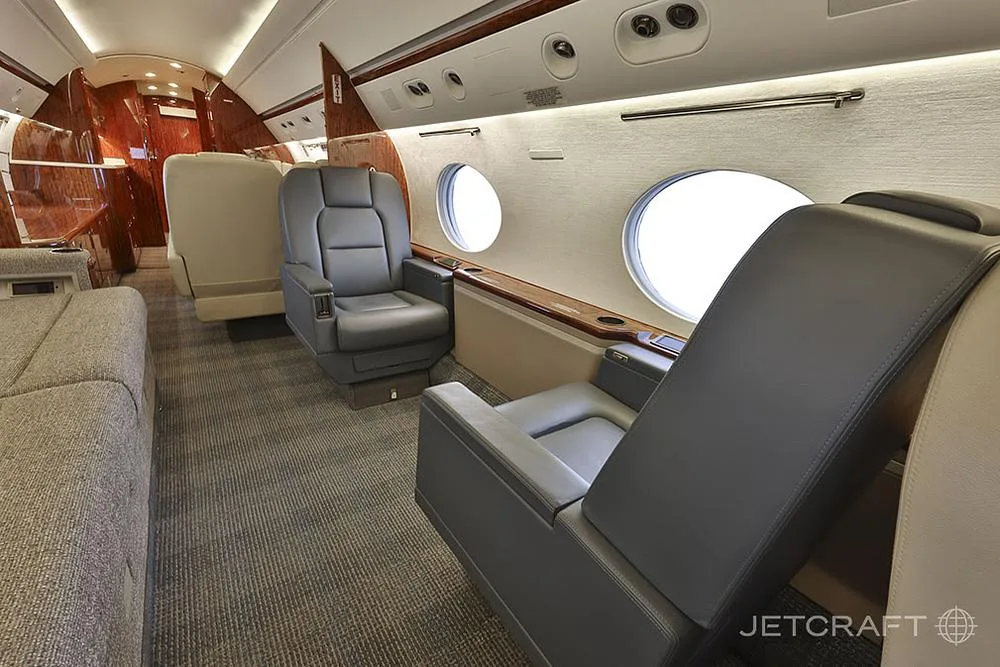 The aft section of the business jet cabin displays individual dark gray leather seats and a light-colored sofa.