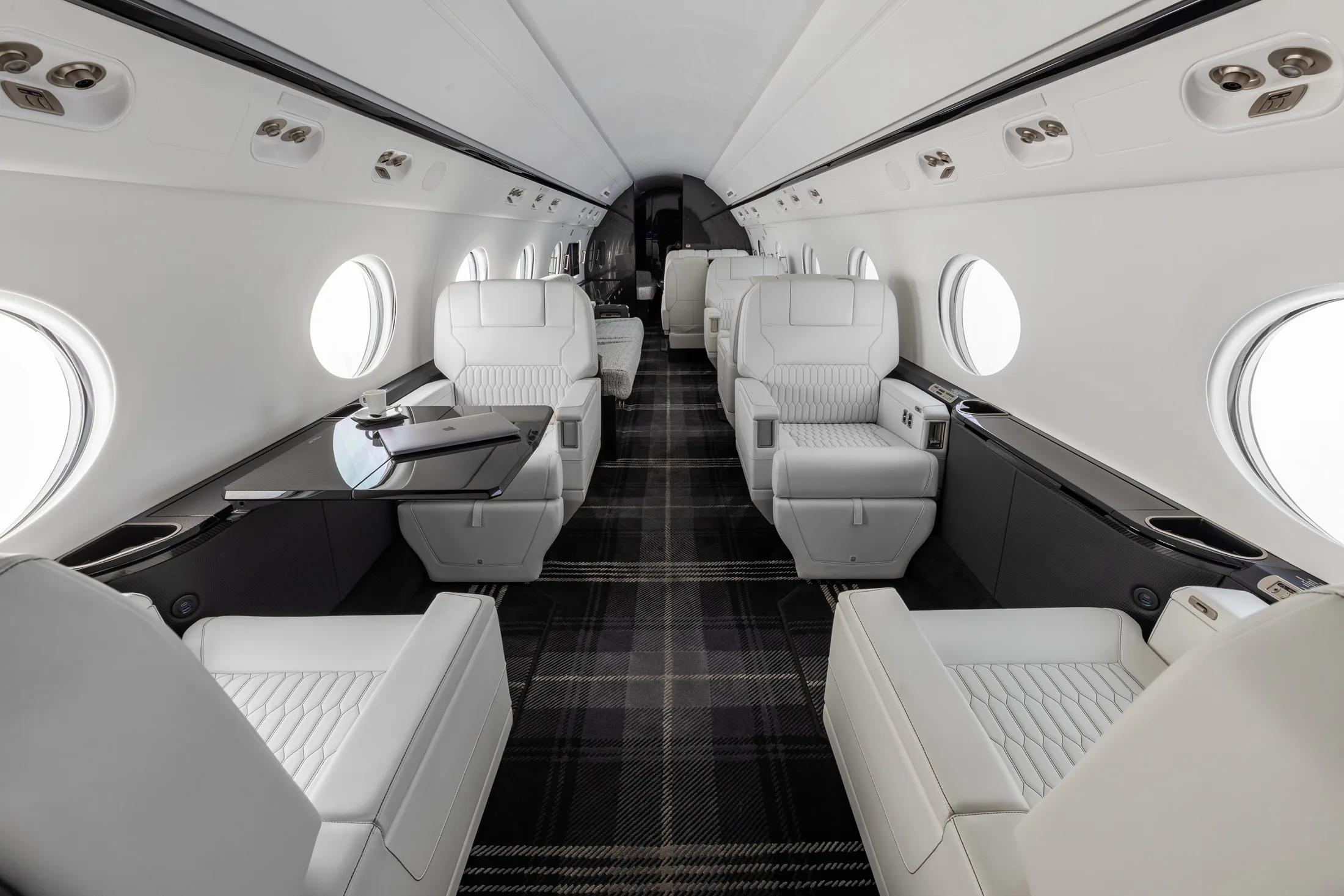 The luxurious interior cabin of a private jet, featuring white leather seats, a dark plaid carpet, and a table with a laptop.
