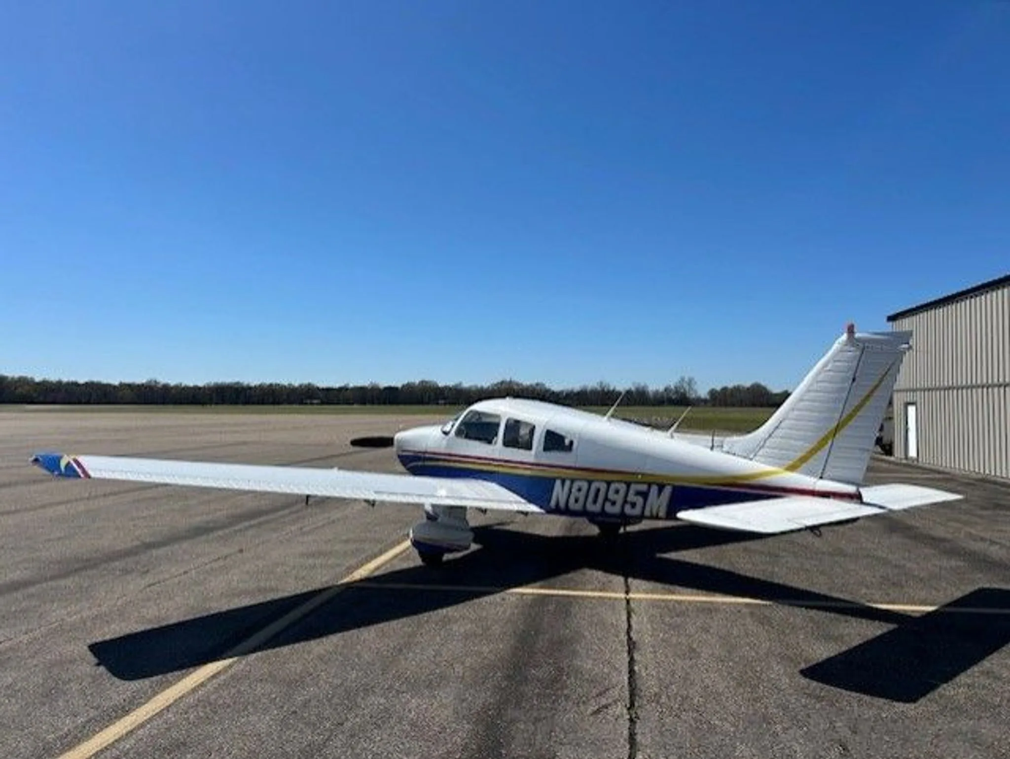 A side view of a white and blue Piper Cherokee aircraft with N8095M registration on an airfield under a clear sky.